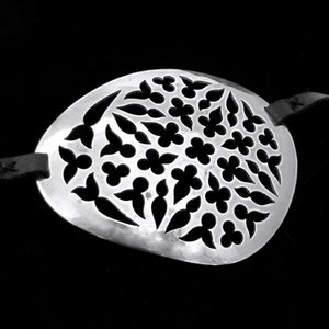 May include: A silver eye patch with a decorative floral pattern cut out. The eye patch is attached to a black leather strap.