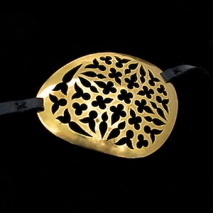 May include: A gold-coloured eye patch with a decorative, intricate, floral pattern. The patch is attached to a black leather strap.