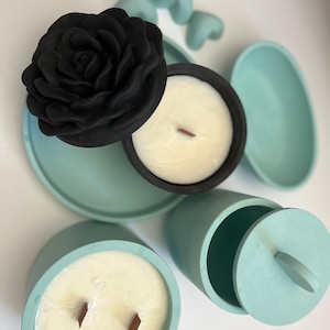Könnte beinhalten: Zwei tealblaue Kerzenbehälter mit Deckeln. Ein Behälter hat einen schwarzen Rosen-Deckel und eine weiße Kerze mit einem Holzdocht. Der andere Behälter hat eine weiße Kerze mit zwei Holzdochten.