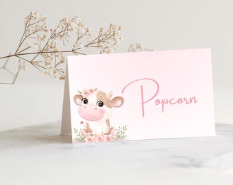 May include: A pink card with the word "Popcorn" in cursive script. The card features a cartoon cow with a floral crown and pink accents. The cow is surrounded by pink flowers and green leaves.