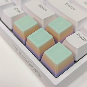 May include: Four pastel-colored keycaps with a gradient design. The keycaps are light green, yellow, orange, and purple. The keycaps are on a white keyboard.