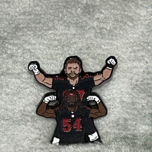 May include: A black and red enamel pin featuring two football players. The top player wears the number 57, and the bottom player wears the number 54. Both are in a celebratory pose with their arms raised.