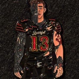 May include: A black and red enamel pin depicting a football player with a cyborg half-face and arm. The jersey displays the number 13 in red and gold, with the word "Faithful" above. The pin is set against a dark backdrop.