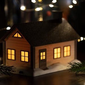 Może przedstawiać: Miniaturowa figurka domku z ciemnym dachem i jasnymi ścianami, posiadająca podświetlane okna oraz mały komin. Konstrukcja stoi na białej podstawie przypominającej śnieg, a z wnętrza okien wydobywa się ciepłe, żółte światło.
