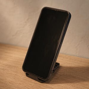 May include: A dark-coloured smartphone with a black screen is positioned upright on a black adjustable stand. The device features a slim frame and is angled slightly to the right, showcasing the side buttons and the sleek, minimalist design of the holder.