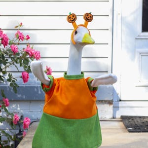 May include: A goose wearing a Halloween costume. The costume includes an orange and green dress, a headband with pumpkin decorations, and matching green shoes. The goose is standing on a concrete step in front of a white house.