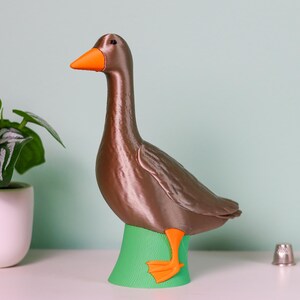 May include: A brown goose figurine with an orange beak and feet stands on a green base. A small potted plant and a spool of thread are also visible. The goose is a decorative item.