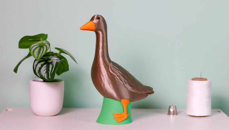May include: A brown goose figurine with an orange beak and feet stands on a green base. A small potted plant and a spool of thread are also visible. The goose is a decorative item.
