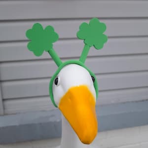 May include: A white goose wearing a green headband with shamrock shaped ears. The headband is made of foam and has a green band that goes around the goose's head.