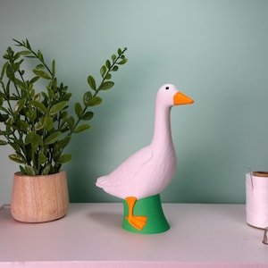 May include: A white goose figurine with an orange beak and feet, perched on a green base. A potted plant and a spool of thread are also visible. The figurine is a decorative item.