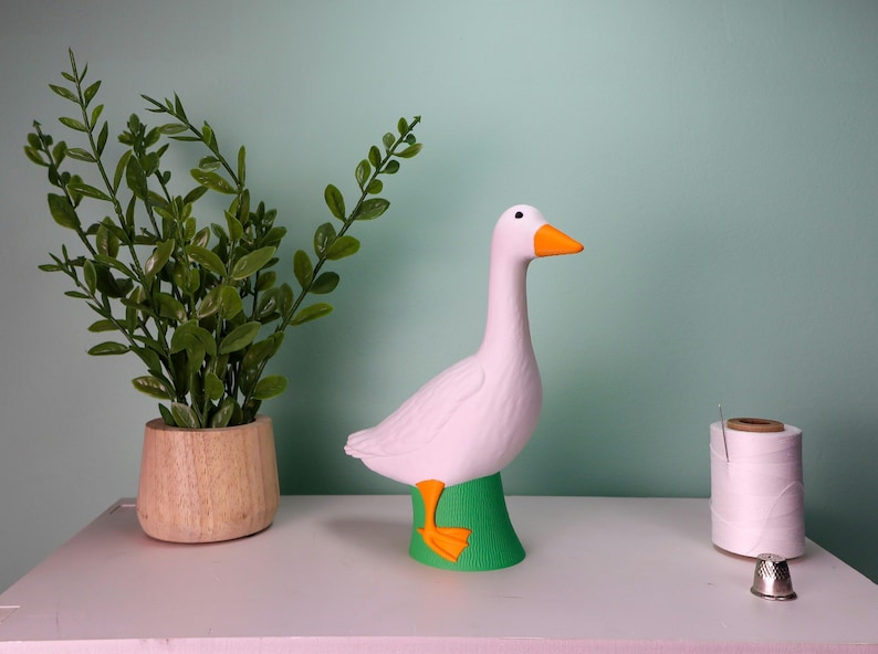 May include: A white goose figurine with an orange beak and feet, perched on a green base. A potted plant and a spool of thread are also visible. The figurine is a decorative item.