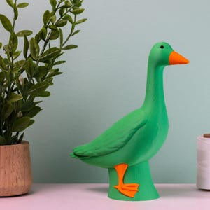 May include: A vibrant green goose figurine with an orange beak and feet. The decorative goose is standing on a green base. A small potted plant and sewing supplies are in the background.