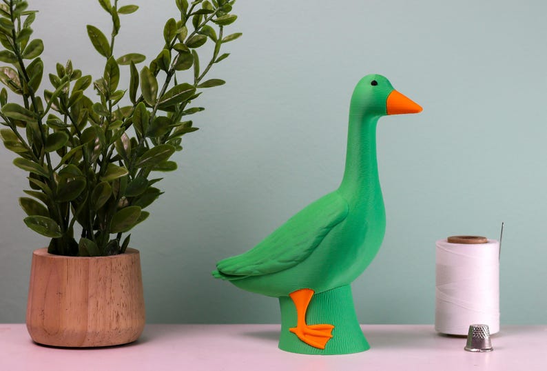 May include: A vibrant green goose figurine with an orange beak and feet. The decorative goose is standing on a green base. A small potted plant and sewing supplies are in the background.