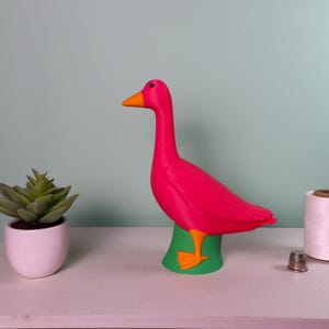 May include: A vibrant pink goose figurine with an orange beak and feet, perched on a green base. A small white potted succulent, a spool of white thread, and a thimble are also on the white surface.