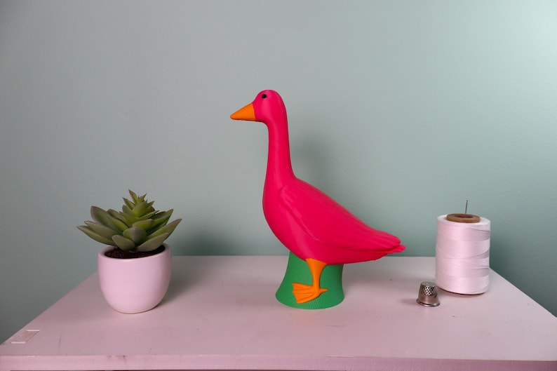 May include: A vibrant pink goose figurine with an orange beak and feet, perched on a green base. A small white potted succulent, a spool of white thread, and a thimble are also on the white surface.