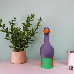 May include: A whimsical purple goose figurine with an orange beak and feet, and a green base. A small potted plant and sewing supplies are also on the table. The goose is a decorative item.