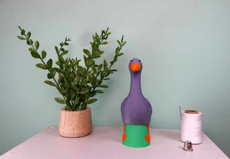 May include: A whimsical purple goose figurine with an orange beak and feet, and a green base. A small potted plant and sewing supplies are also on the table. The goose is a decorative item.