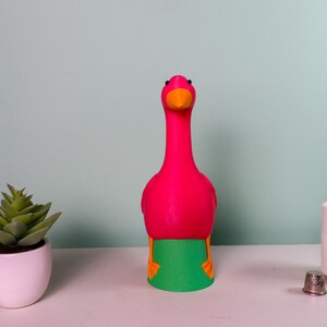 May include: A vibrant pink flamingo-shaped decorative item with an orange beak and green and orange accents. A small potted succulent, a spool of white thread, and a thimble are also on the white surface.