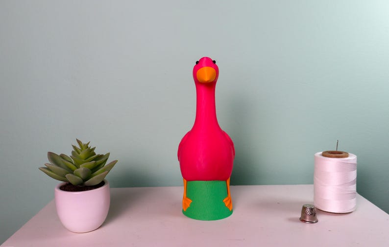 May include: A vibrant pink flamingo-shaped decorative item with an orange beak and green and orange accents. A small potted succulent, a spool of white thread, and a thimble are also on the white surface.