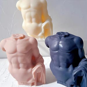 Pode incluir: Três esculturas em forma de torso em cores variadas: amarelo pálido, rosa empoeirado e azul marinho. Cada escultura retrata um torso masculino musculoso com tecido drapeado. As esculturas são exibidas em uma superfície branca.