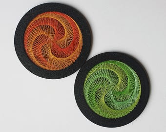 May include: Two circular string art pieces. One has orange and yellow threads, the other green and yellow. Both are in black circular frames. The string art forms a spiral design.