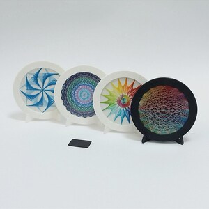 May include: Four circular string art displays on stands. Three have white frames and one has a black frame. The designs feature intricate geometric patterns created with blue, purple, green, yellow, and orange thread. A small black rectangular object sits in front.