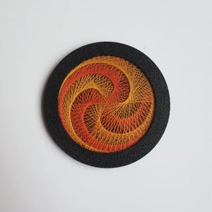 May include: A round, black framed string art piece. The artwork features a spiral design in shades of orange and yellow thread. The threads are intricately woven to create a textured, eye-catching pattern. The piece is set against a white background.