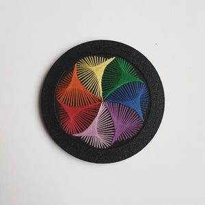 May include: A round, black coaster with a colorful, embroidered design. The design features a rainbow of colors, including red, orange, yellow, green, blue, and purple, arranged in a radial pattern. The coaster is likely made of wood or a similar material.