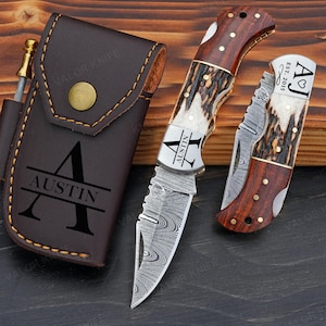 Peut inclure: Un étui à couteau en cuir marron avec le nom "AUSTIN" et un grand "A" est à côté d'un couteau avec un manche en bois et une lame à motifs. Un couteau plus petit avec une conception de manche et de lame similaire est également présent.