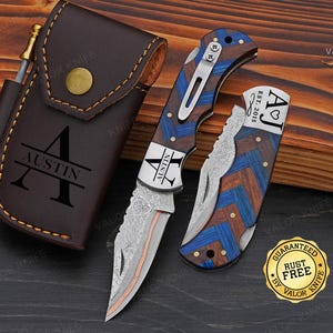 May include: A personalized pocket knife set with a brown leather sheath, a sharpening rod, and two folding knives. The knives feature a wood and blue chevron pattern, with the name "Austin" engraved on the blade. The set includes a "Rust Free" guarantee.