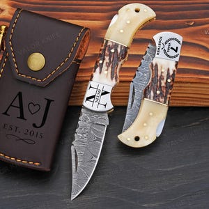 Peut inclure: Un étui à couteau en cuir marron avec un bouton doré et les initiales "A J" et l'année "EST. 2015" gravées. Deux couteaux avec des manches en os et des lames à motifs sont à côté de l'étui. Un couteau est ouvert et l'autre est fermé.