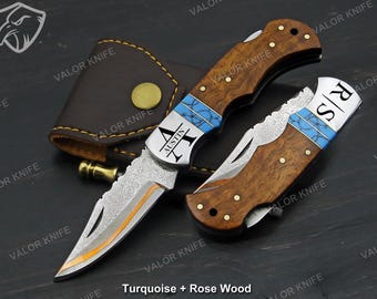 Op de afbeelding: Twee vouwmessen met rozenhouten handvatten en turquoise accenten. Eén mes is open en toont een patroonmes met een goudkleurige rand. Het andere mes is gesloten. De tekst "AUSTIN" en initialen zijn op de messen gegraveerd. Een leren foedraal is zichtbaar.