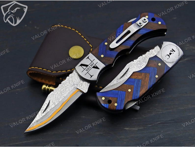 May include: A folding pocket knife with a Damascus steel blade featuring an orange edge, and a handle with a blue and brown chevron pattern. The metal bolster is engraved with the text NELSON IV, and the other handle section features K C.