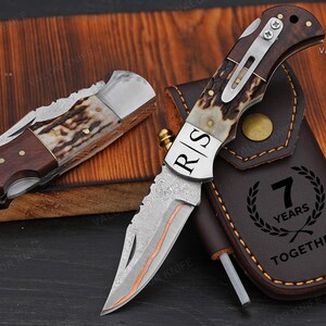 May include: Two folding knives with wood and antler handles, one closed and one open, featuring a Damascus steel blade with the letters R|S engraved. A brown leather sheath is included, embossed with the text 7 YEARS TOGETHER and a laurel wreath.