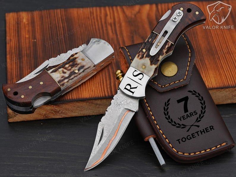 May include: Two folding knives with wood and antler handles, one closed and one open, featuring a Damascus steel blade with the letters R|S engraved. A brown leather sheath is included, embossed with the text 7 YEARS TOGETHER and a laurel wreath.