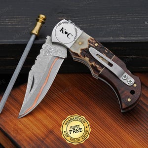 May include: A folding pocket knife with a Damascus steel blade featuring a copper inlay and a shield emblem. The handle is made of brown wood and textured antler with a silver pocket clip. The bolster is engraved with K C and a heart symbol.