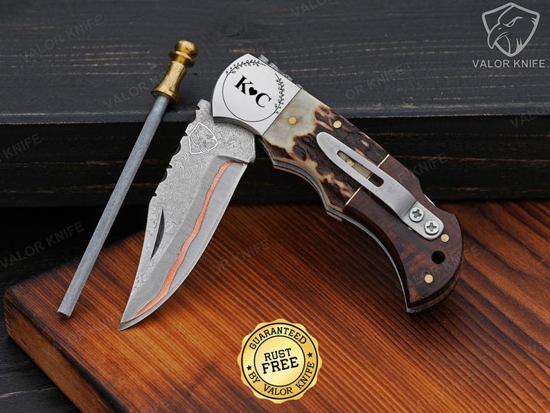 May include: A folding pocket knife with a Damascus steel blade featuring a copper inlay and a shield emblem. The handle is made of brown wood and textured antler with a silver pocket clip. The bolster is engraved with K C and a heart symbol.