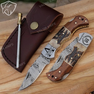 May include: A personalized pocket knife with a Damascus steel blade, a stag horn handle, and a wooden inlay. The knife is engraved with the name "Arther" and the text "Together 10 Years". It comes with a brown leather sheath.