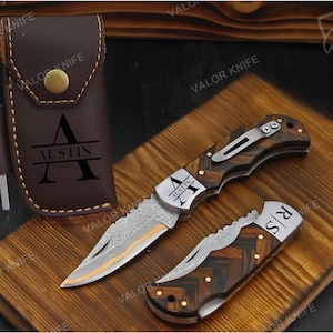 May include: A brown leather sheath with a brass snap and the name AUSTIN engraved, paired with two folding knives featuring Damascus steel blades and wood handles. One knife is open, one is closed, and both feature personalized engravings.