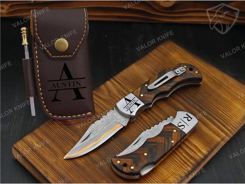 May include: A brown leather sheath with a brass snap and the name AUSTIN engraved, paired with two folding knives featuring Damascus steel blades and wood handles. One knife is open, one is closed, and both feature personalized engravings.