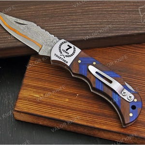 May include: A folding pocket knife with a Damascus steel blade featuring an orange spine accent. The handle is crafted from dark wood with blue chevron inlays and a silver metal clip. A silver bolster displays the text 7 YEARS TOGETHER.
