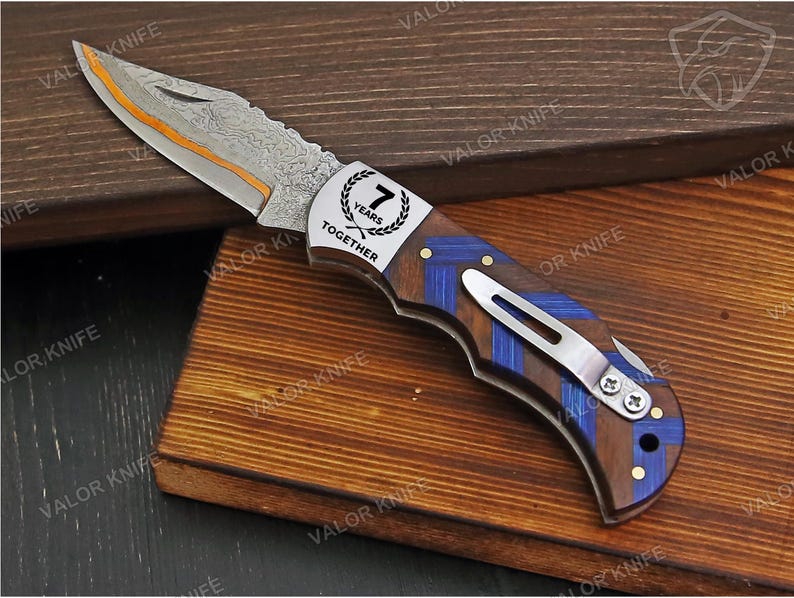 May include: A folding pocket knife with a Damascus steel blade featuring an orange spine accent. The handle is crafted from dark wood with blue chevron inlays and a silver metal clip. A silver bolster displays the text 7 YEARS TOGETHER.