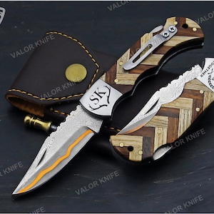 May include: A folding pocket knife with a Damascus steel blade featuring an orange accent line. The handle has a herringbone pattern of light and dark wood. A metal bolster displays the letters SK and a deer antler icon, while the other end shows 7 YEARS TOGETHER.