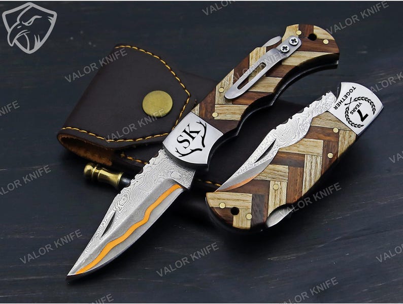 May include: A folding pocket knife with a Damascus steel blade featuring an orange accent line. The handle has a herringbone pattern of light and dark wood. A metal bolster displays the letters SK and a deer antler icon, while the other end shows 7 YEARS TOGETHER.