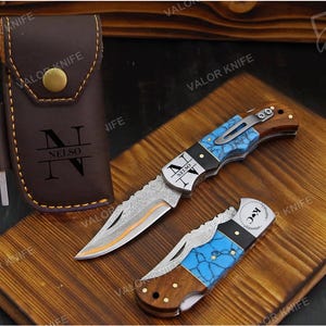 May include: A brown leather sheath with a brass snap and the text "NELSO" features a side loop holding a small metal tool. Two folding pocket knives with blue turquoise-patterned handles, wood accents, and patterned metal blades are displayed.