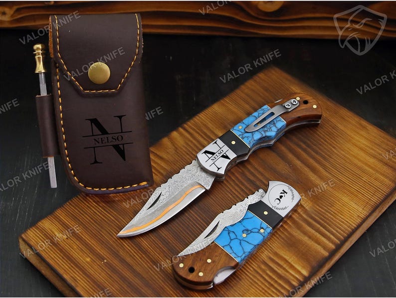 May include: A brown leather sheath with a brass snap and the text "NELSO" features a side loop holding a small metal tool. Two folding pocket knives with blue turquoise-patterned handles, wood accents, and patterned metal blades are displayed.
