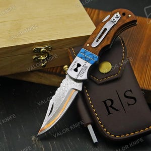 May include: A folding pocket knife with a wood handle, a silver metal pocket clip, and a patterned blade. The handle features a blue inlay and is engraved with "MASON GROOMSMAN 12.10.2020" and a bowtie icon. A brown leather sheath with "R|S" is included.