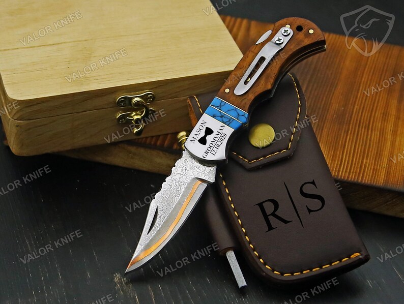 May include: A folding pocket knife with a wood handle, a silver metal pocket clip, and a patterned blade. The handle features a blue inlay and is engraved with "MASON GROOMSMAN 12.10.2020" and a bowtie icon. A brown leather sheath with "R|S" is included.