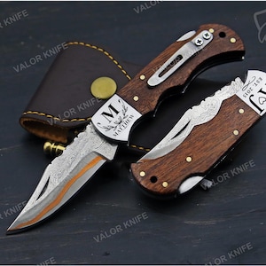 May include: Two folding pocket knives with dark wood handles and metal bolsters. One knife is open, showing a patterned blade with an orange accent. The bolster is engraved with 'M MATTHEW'. The closed knife features 'AJ EST. 2015'. A brown leather sheath is included.