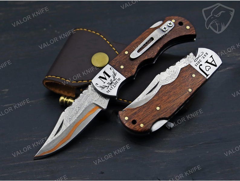 May include: Two folding pocket knives with dark wood handles and metal bolsters. One knife is open, showing a patterned blade with an orange accent. The bolster is engraved with 'M MATTHEW'. The closed knife features 'AJ EST. 2015'. A brown leather sheath is included.